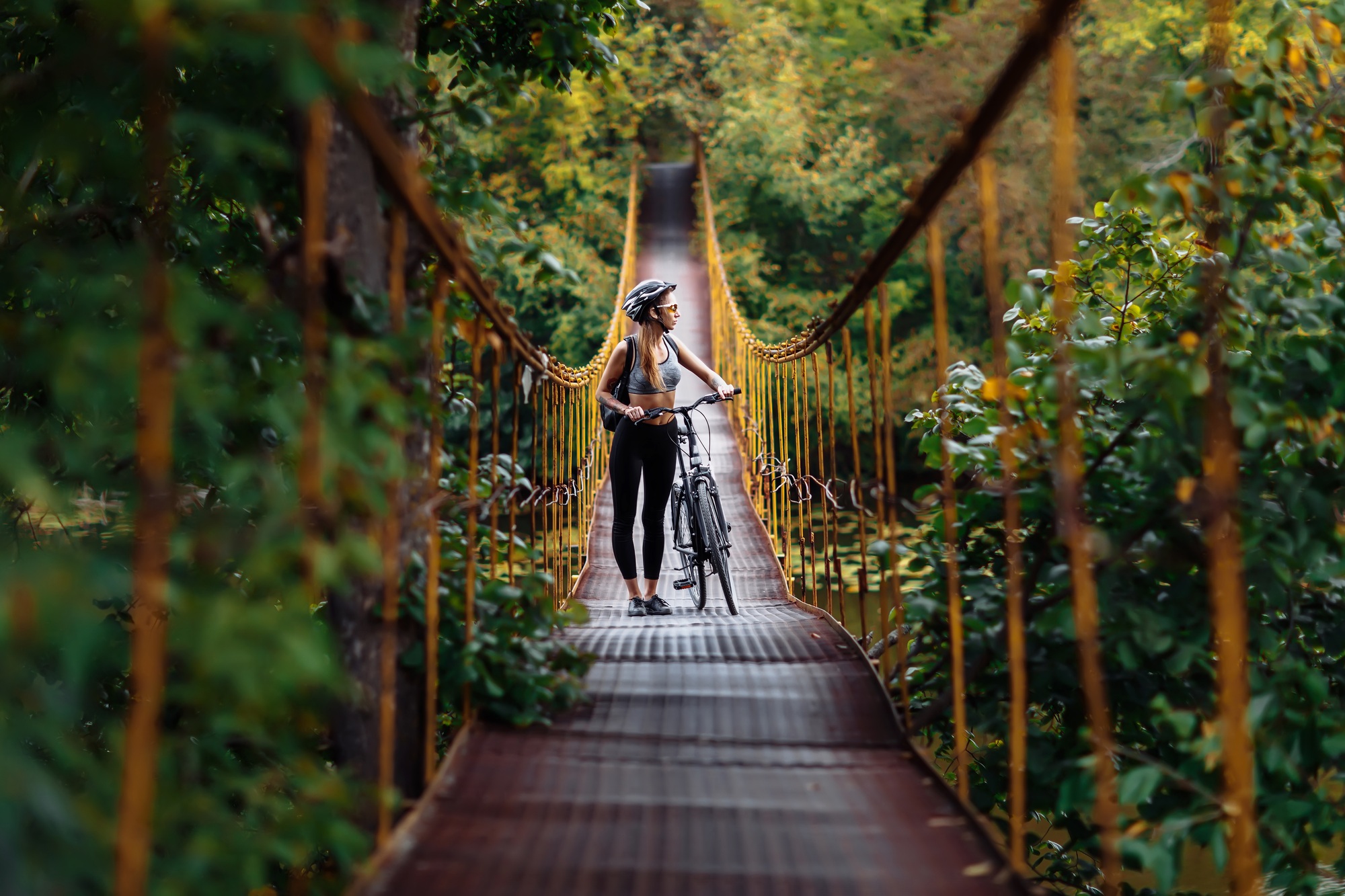 Fit young woman wearing sportswear with her bicycle on suspension bridge at autumn